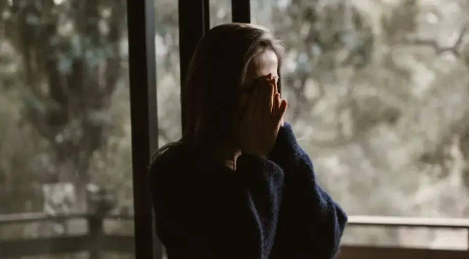 A woman standing by a window with her hands covering her face, appearing overwhelmed or sorrowful, with a blurred forest background suggesting a secluded or private moment of grief.