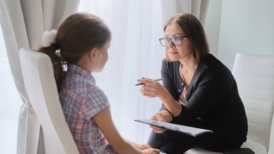 A female therapist and a young girl, both Caucasian, sit facing each other in a bright, modern office. The therapist is holding a pen and notepad, gesturing while speaking to the girl, who looks attentive
