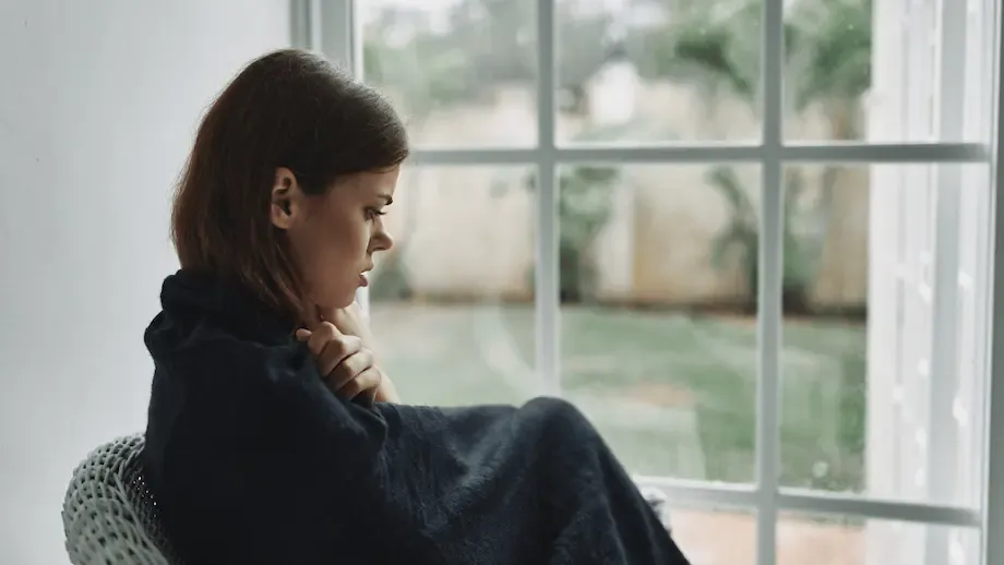 A young woman sitting alone on a wicker chair by a large window, wrapped in a dark blue blanket. She looks thoughtfully out the window with a somber expression, in a room with minimalistic decor and a view of a garden outside