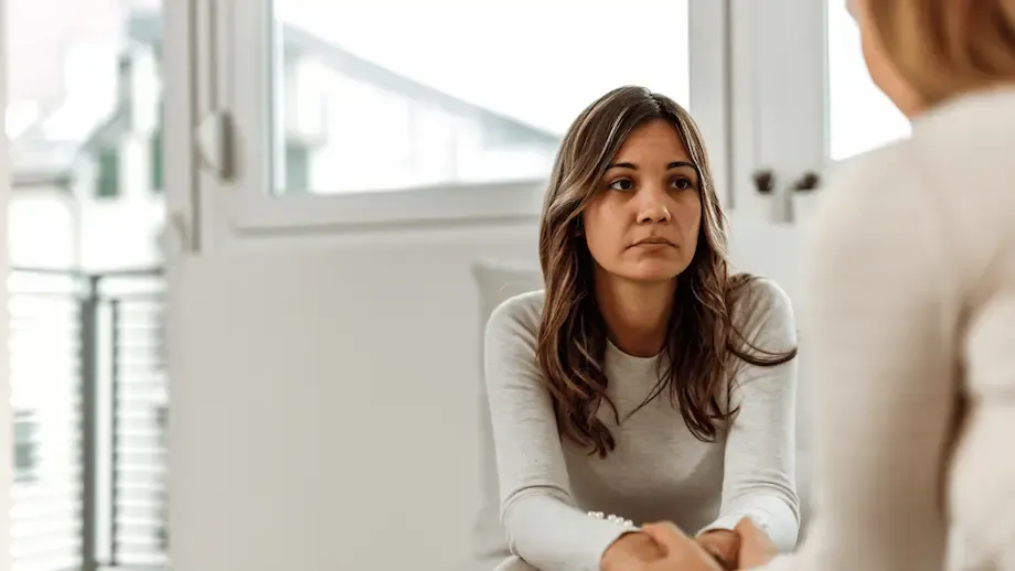 A woman, appearing Hispanic, in a serious conversation with a therapist in a bright and airy room. The woman looks concerned and is seated with her hands clasped together, focusing intently on the therapist.
