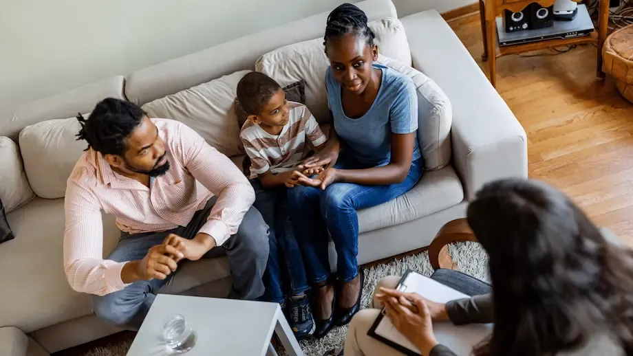 A family therapy session showing a Black family with a father, mother, and young son sitting on a sofa, talking to a female therapist who sits across from them with a notepad